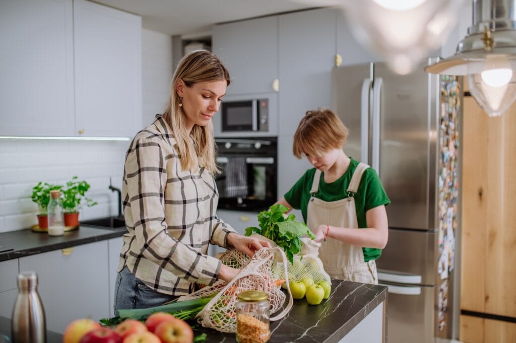 Zero-Waste-Kochen – Nachhaltig & clever: So reduzierst du Küchenabfälle auf ein Minimum 2 - Küchenhelferlein Mother unpacking local food in zero waste packaging from bag with help of daughter in kitchen at